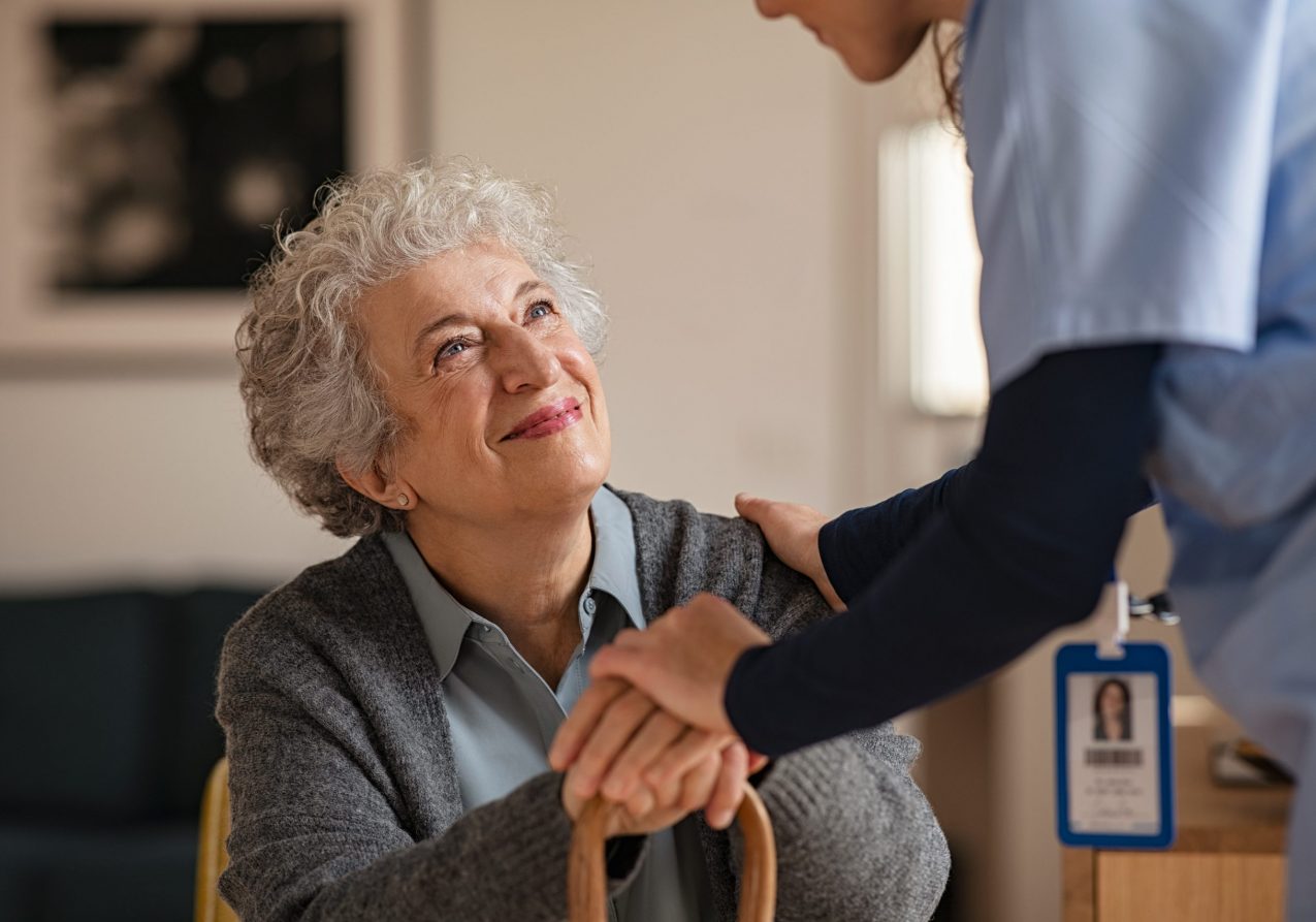 Old woman talking with a female doctor while holding hands at home. Smiling senior woman talking to her general practitioner visiting her at home during virus epidemic. Happy old patient holding hands of caregiver at nursing home.
